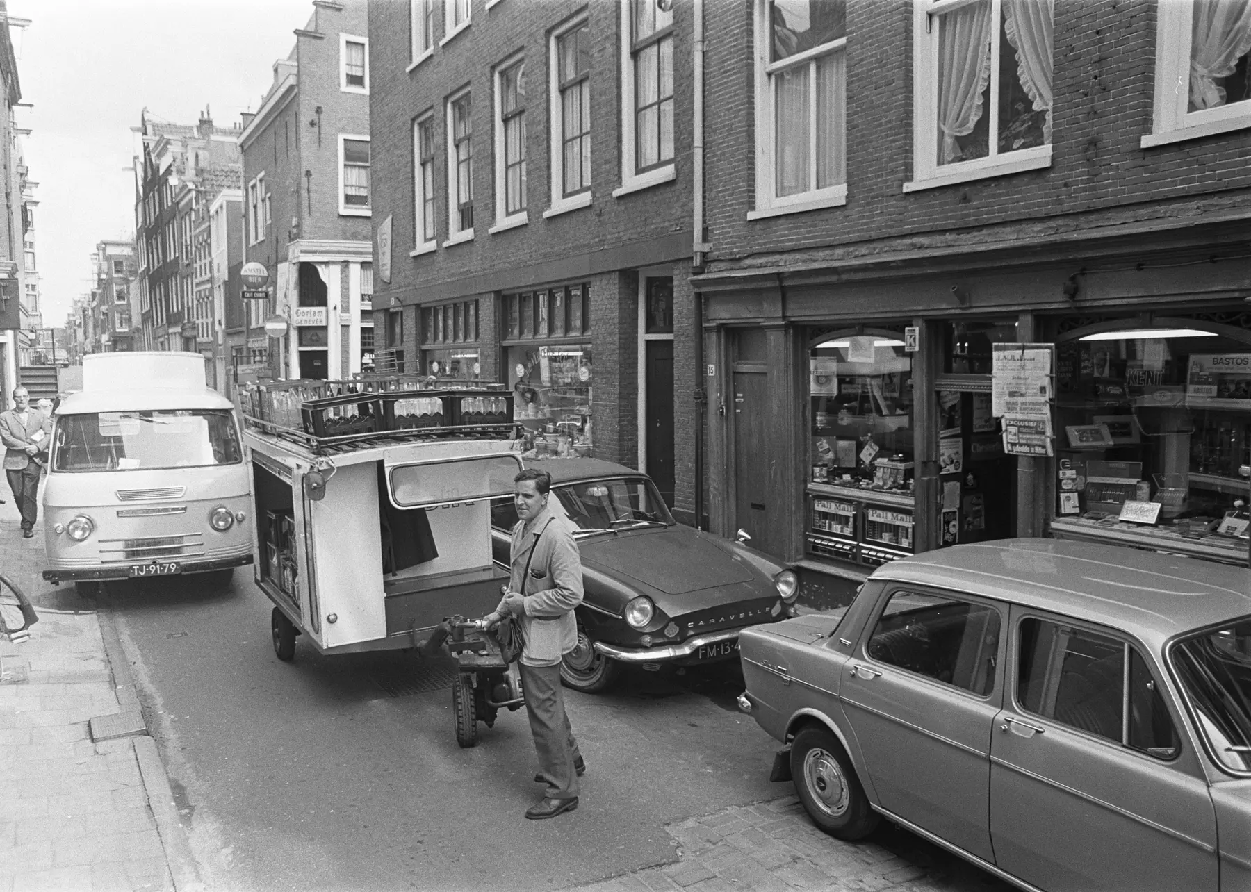 Een melkboer staat in 1967 met zijn gemotoriseerde melkwagen tussen auto's in een smalle Nederlandse straat